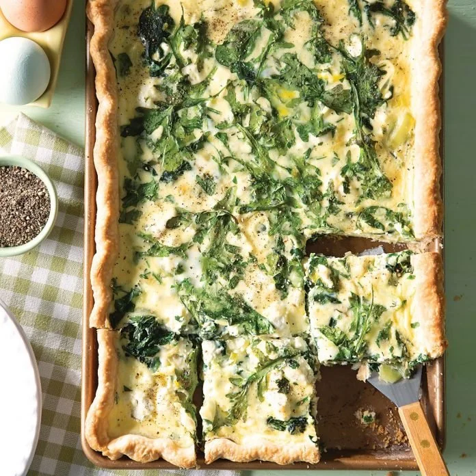 Baked quiche with a golden crust and green leafy vegetables, cut into slices, on a baking dish.