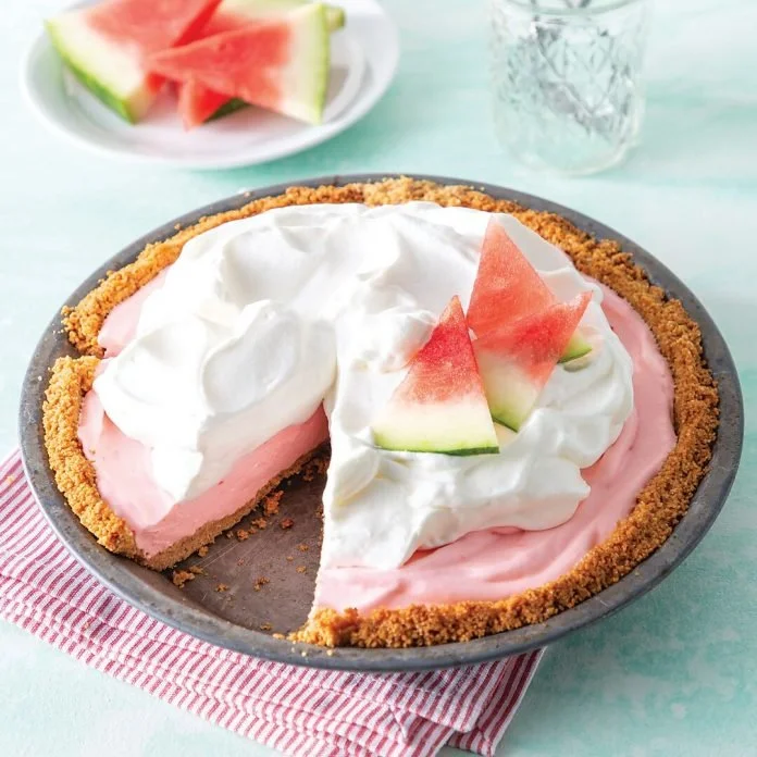 A watermelon-flavored pie with a graham cracker crust, topped with whipped cream and garnished with watermelon slices, on a plate with a red and white striped cloth.