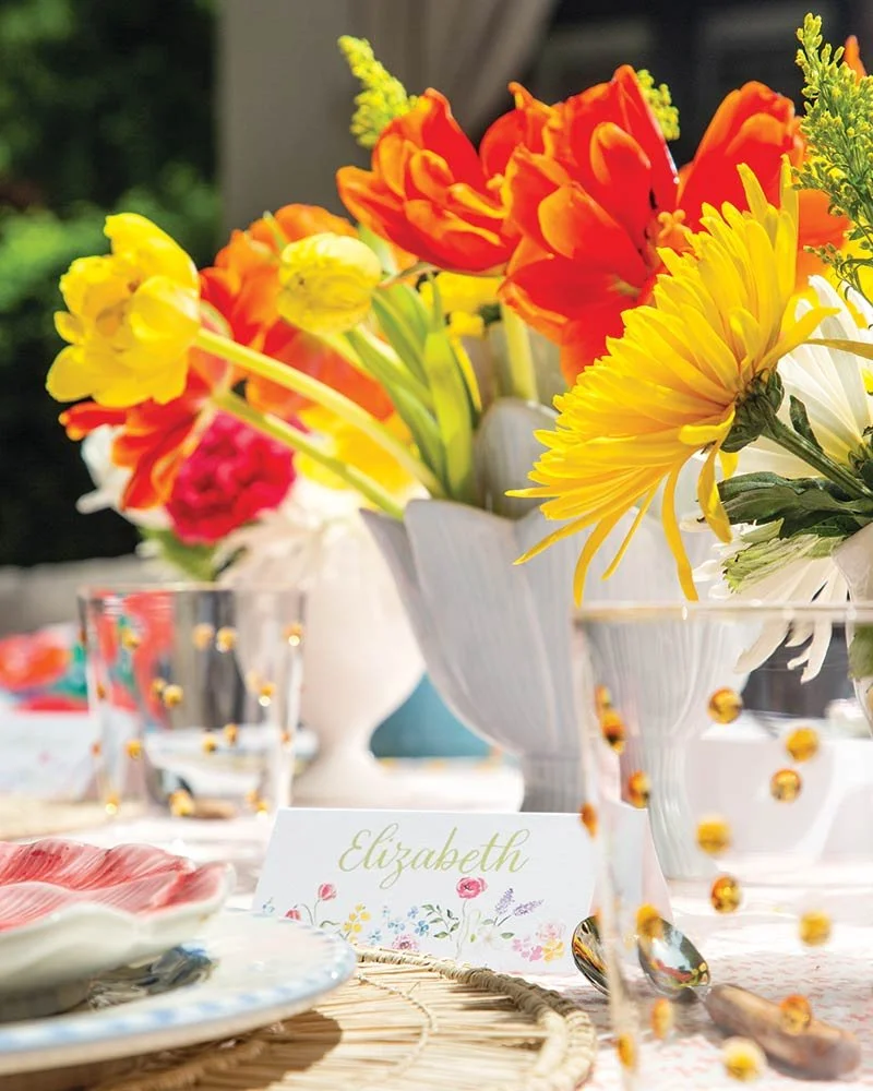 A table setting with vibrant flower arrangements in vases, a place card with the name Elizabeth, and decorative items on a woven placemat.