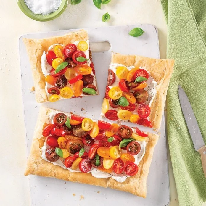 A tray of rectangular flatbread topped with creamy spread, colorful cherry tomatoes, and fresh basil leaves, with a green cloth and a knife nearby.