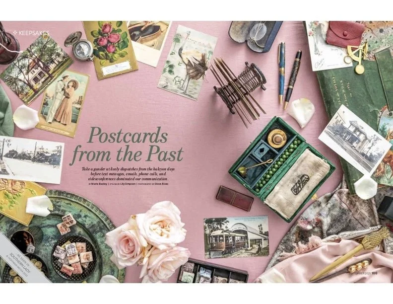 A collection of vintage postcards, photographs, and stationery items arranged on a pink surface, with some flowers, pens, and decorative objects.