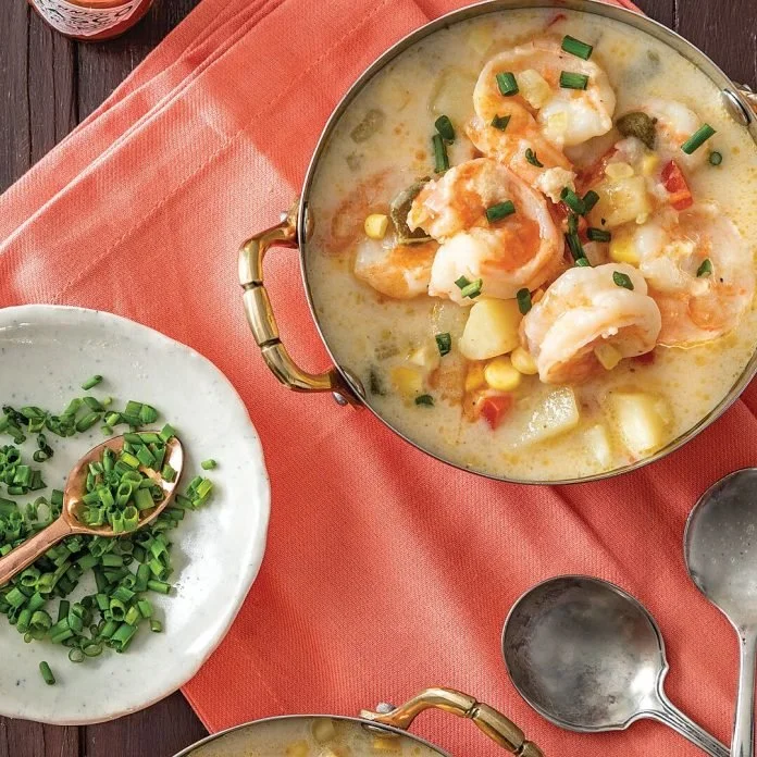 Bowl of creamy shrimp and vegetable chowder garnished with chopped chives, served on a pink napkin with empty spoons nearby.