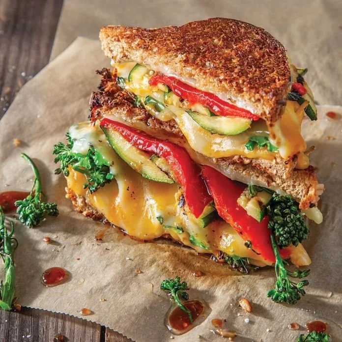A grilled sandwich with melted cheese, red and green bell peppers, cucumbers, and fresh parsley, served on parchment paper with some drops of sauce and pepper around.