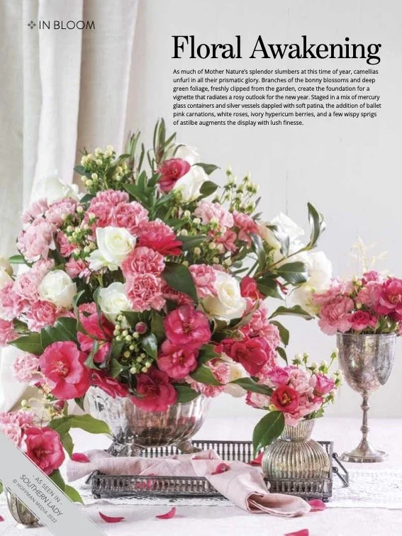 Large floral arrangement with pink and white flowers, green foliage in a clear glass bowl, with a smaller bouquet in a silver vase and a silver goblet on a white tablecloth. Pink petals scattered on the table.