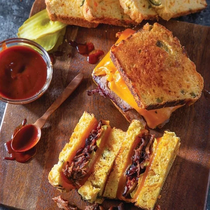 Close-up of a barbecue sandwich with sliced bread, melted cheese, and barbecue beef, served on a wooden cutting board with barbecue sauce on the side and a told of sliced bread with herbs.