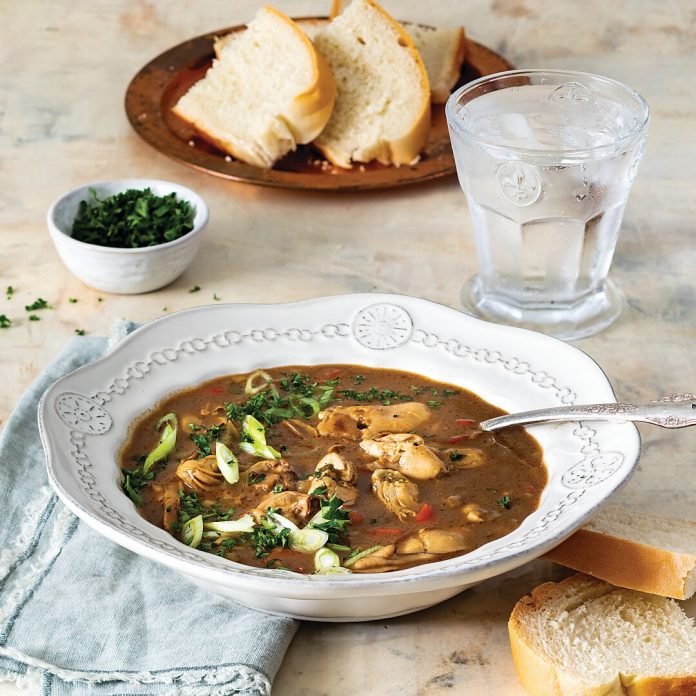 A bowl of chicken noodle soup garnished with chopped green onions and herbs, served with slices of bread, a glass of water with ice, and an additional small bowl of chopped herbs on a light-colored wooden table.