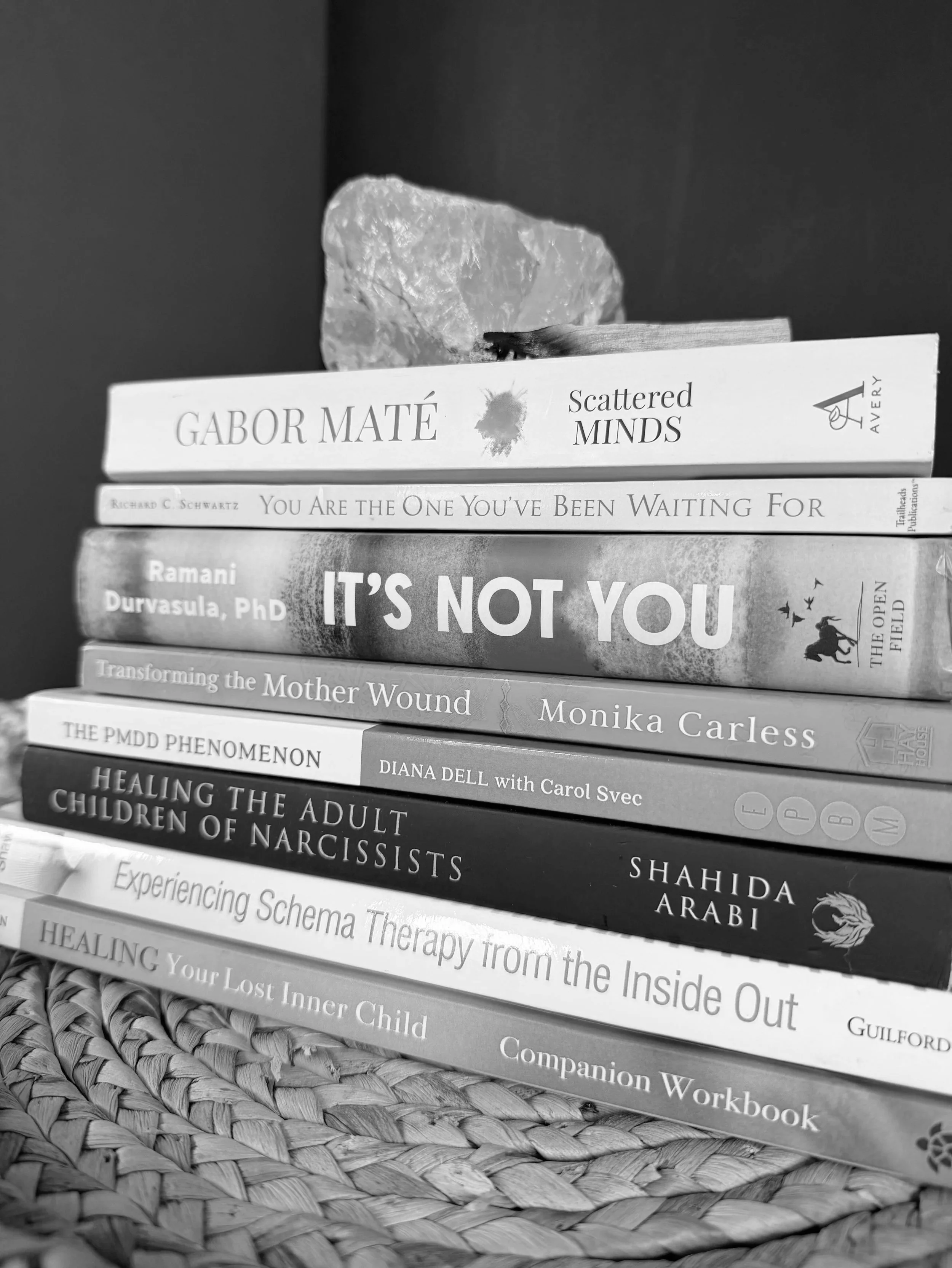 Stack of eight books on a woven surface with a crystal and small twig on top of the books.