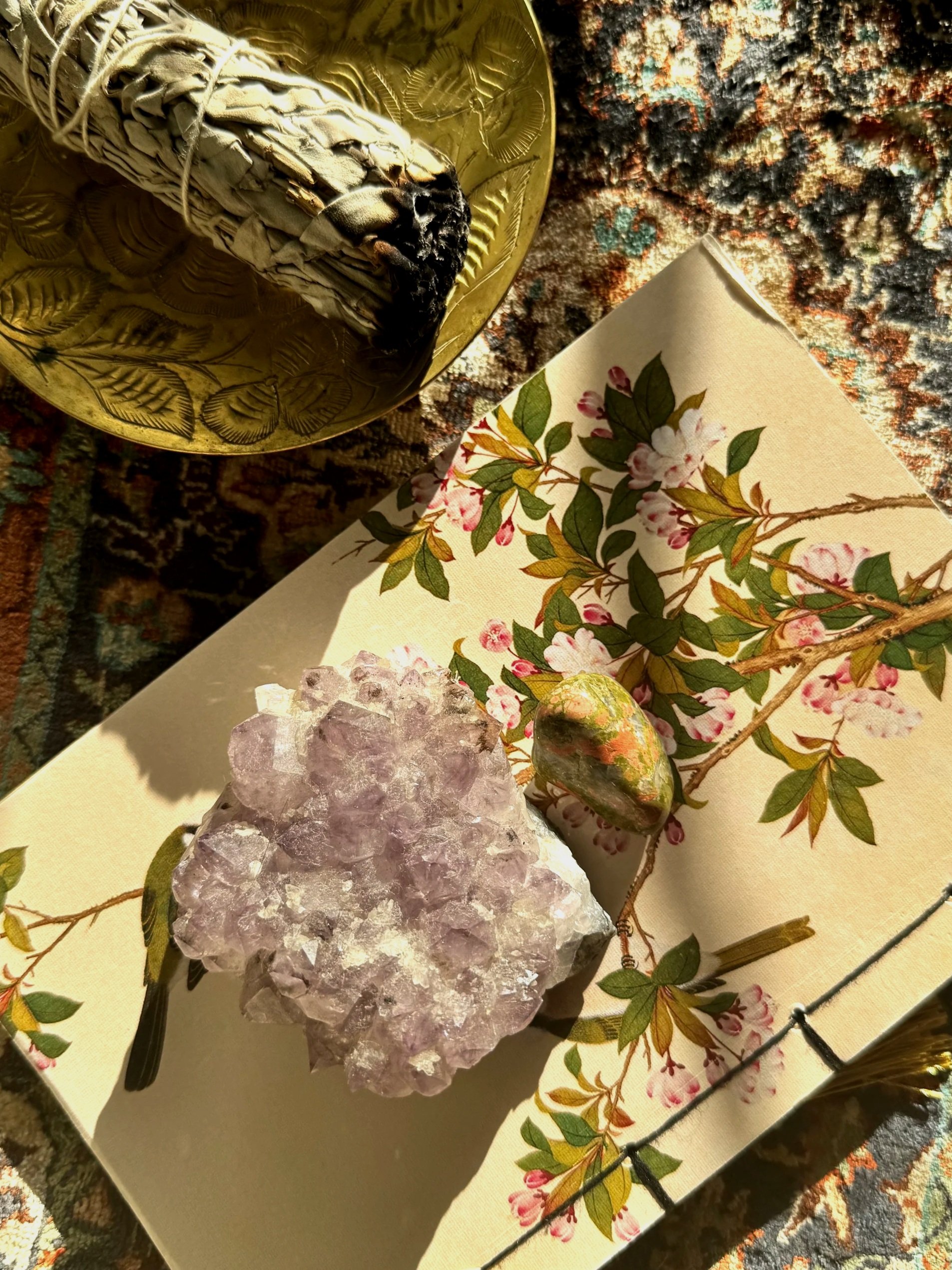 A decorative arrangement featuring a purple mineral cluster, a small multicolored stone, a branch with pink flowers, and a bird figurine on a floral-patterned paper, with a gold decorative dish and a colorful rug in the background.