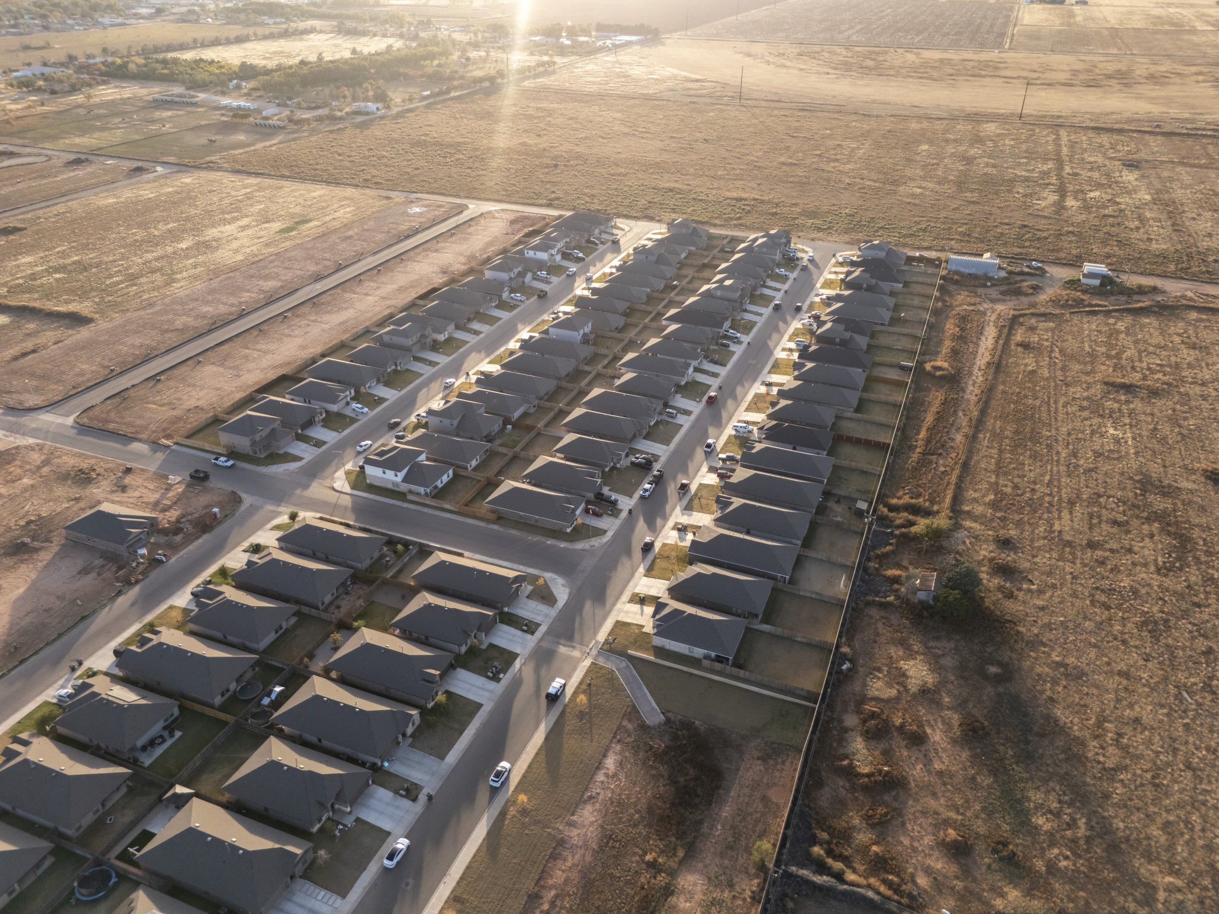 Aerial view of a suburban neighborhood with rows of single-family homes, streets, and parked cars, surrounded by open farmland at sunset.