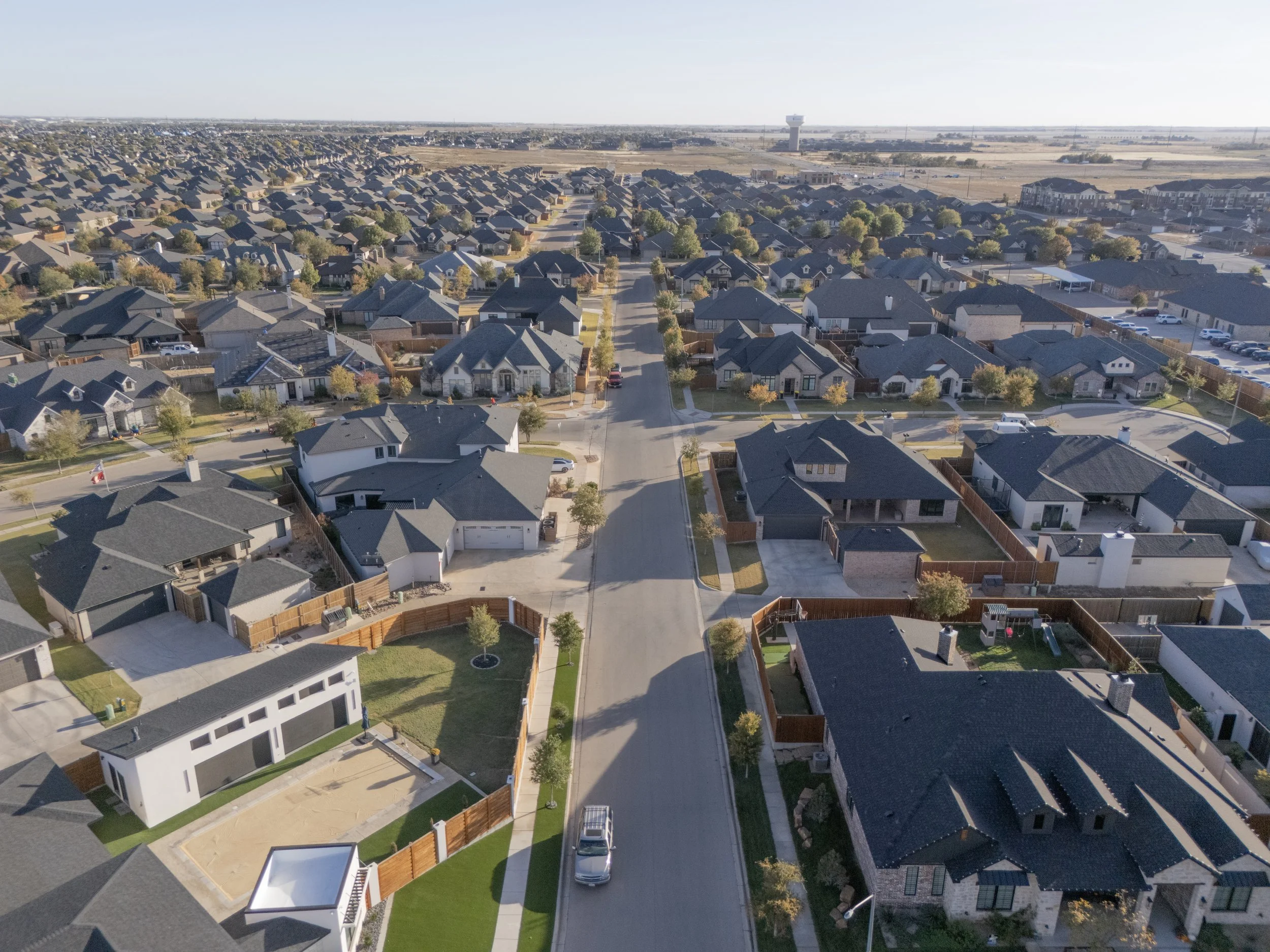 Aerial view of a suburban neighborhood with numerous houses, streets, trees, and cars, with a water tower and open fields in the background.