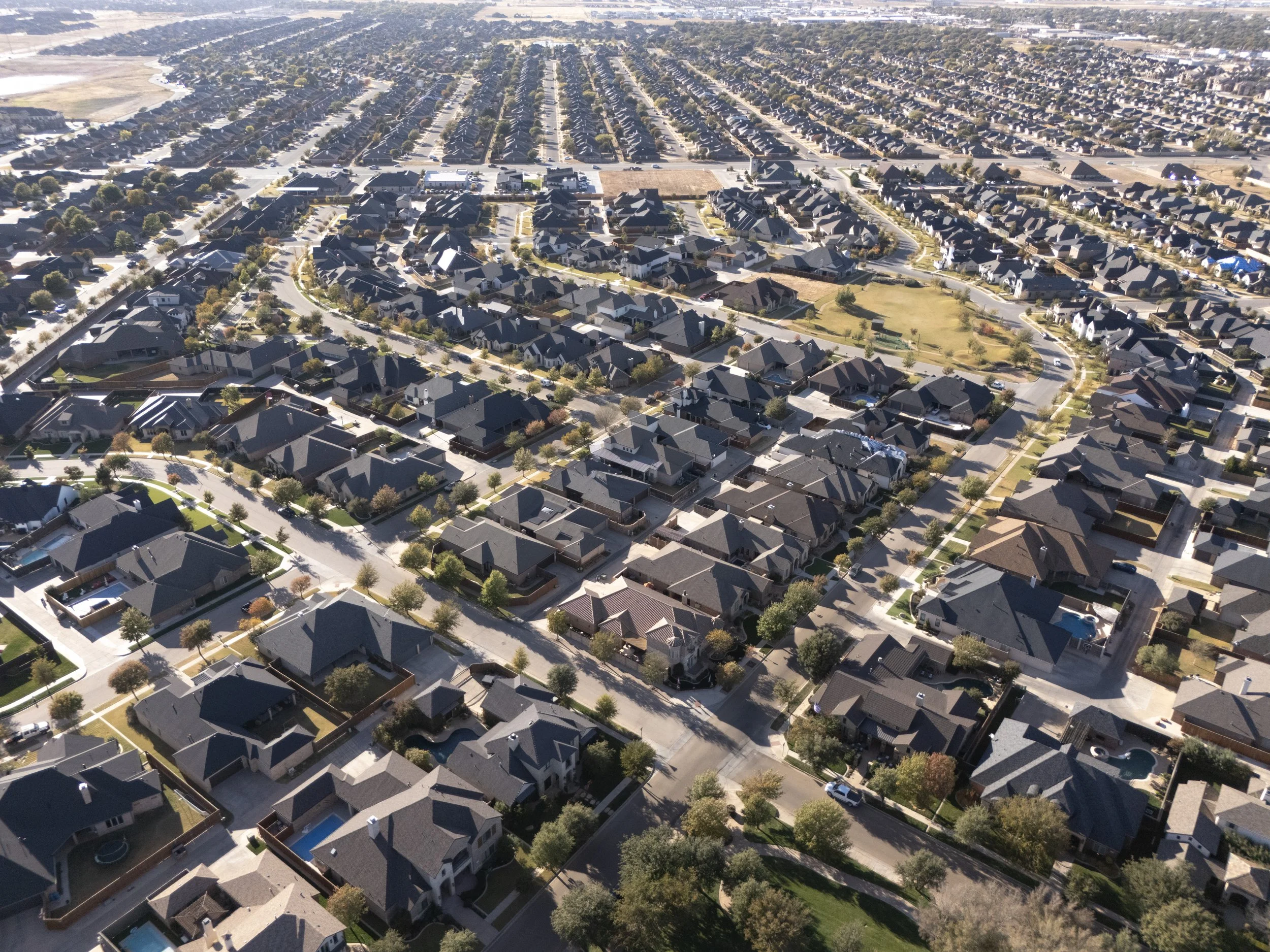 An aerial view of a residential neighborhood with numerous houses, trees lining the streets, and some houses featuring swimming pools in backyards.