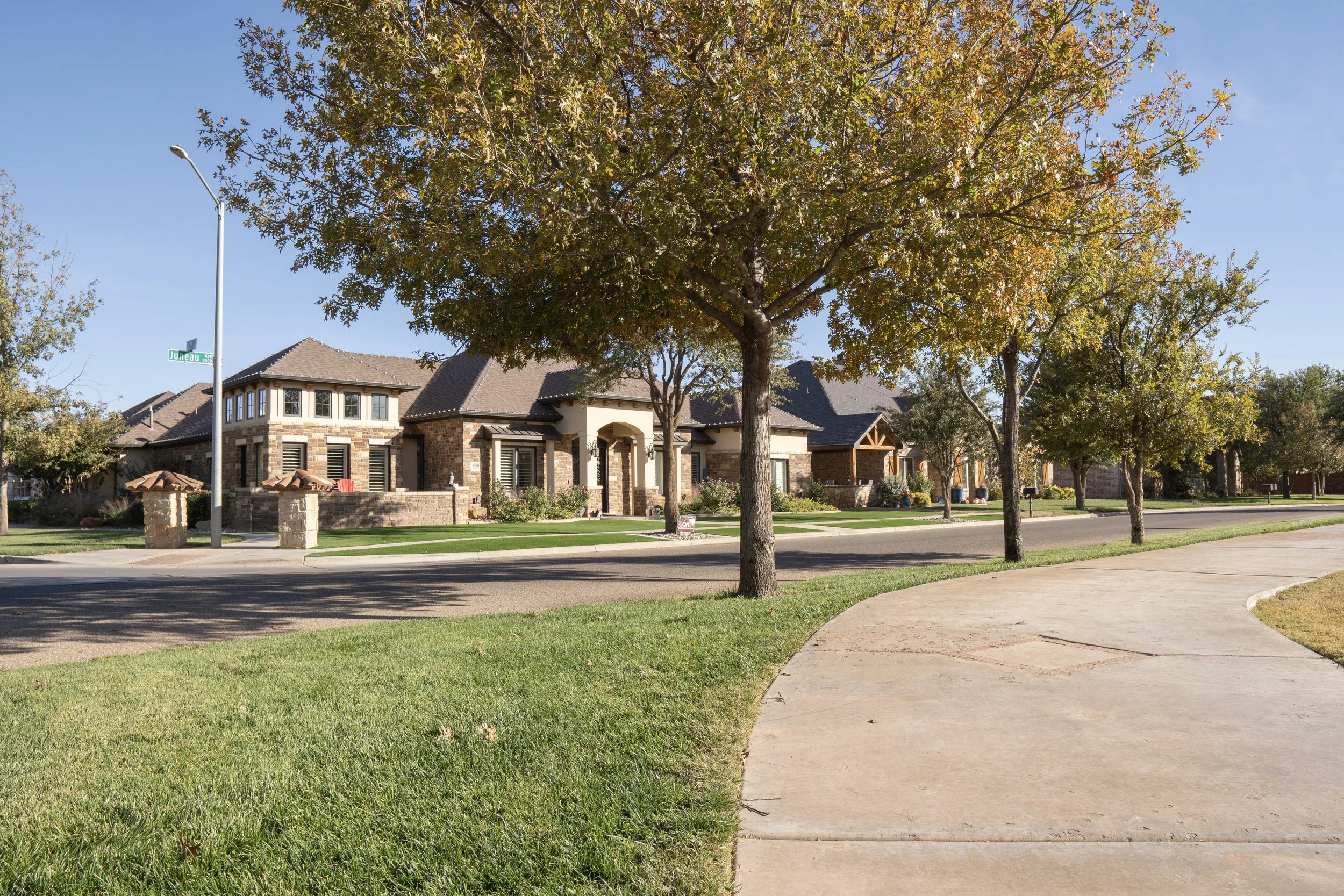 A suburban neighborhood with brick houses, trees lining the streets, a sidewalk, and a clear blue sky.