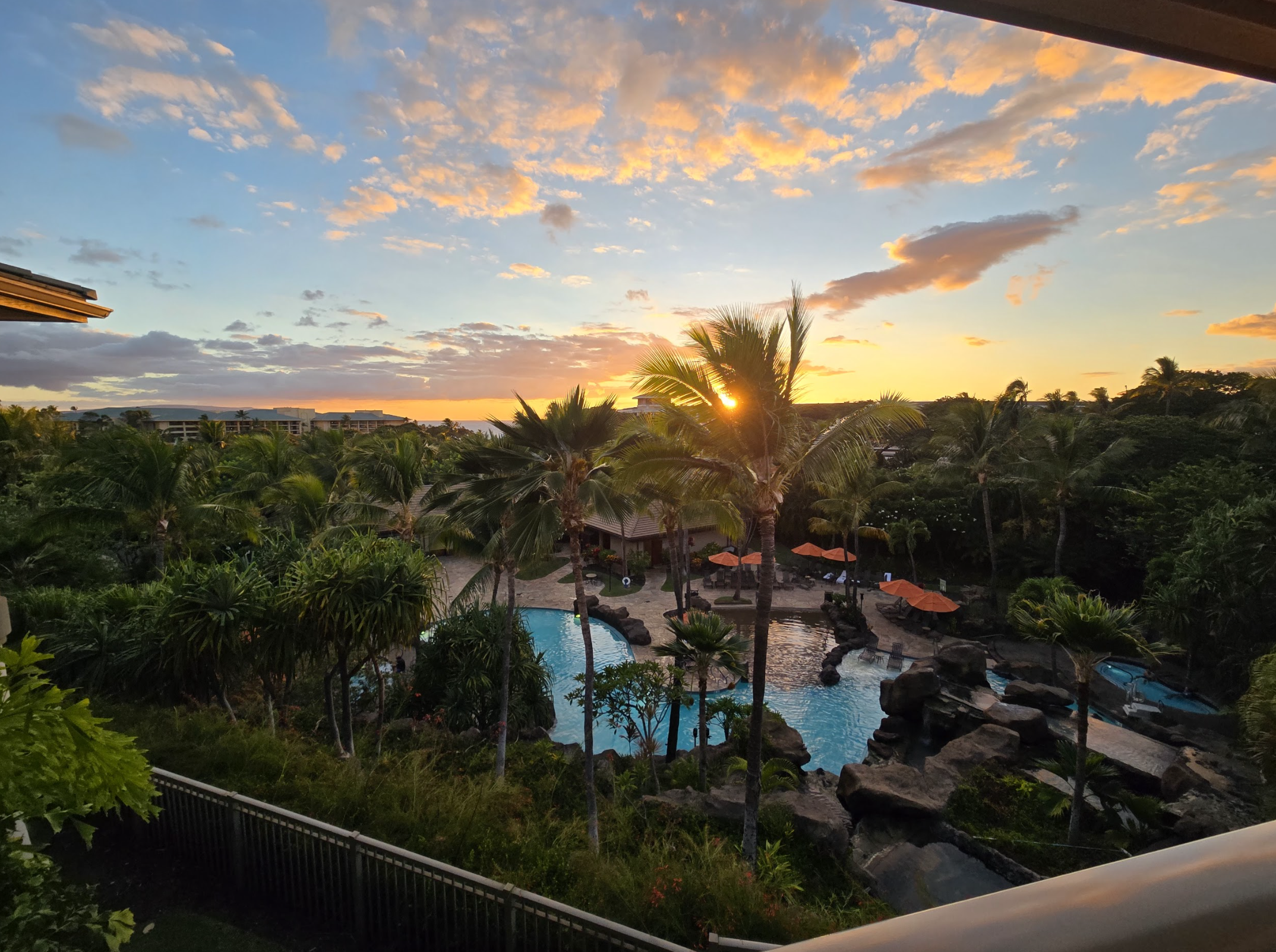 Sunset over a tropical resort with a swimming pool, palm trees, and lounge chairs with umbrellas.
