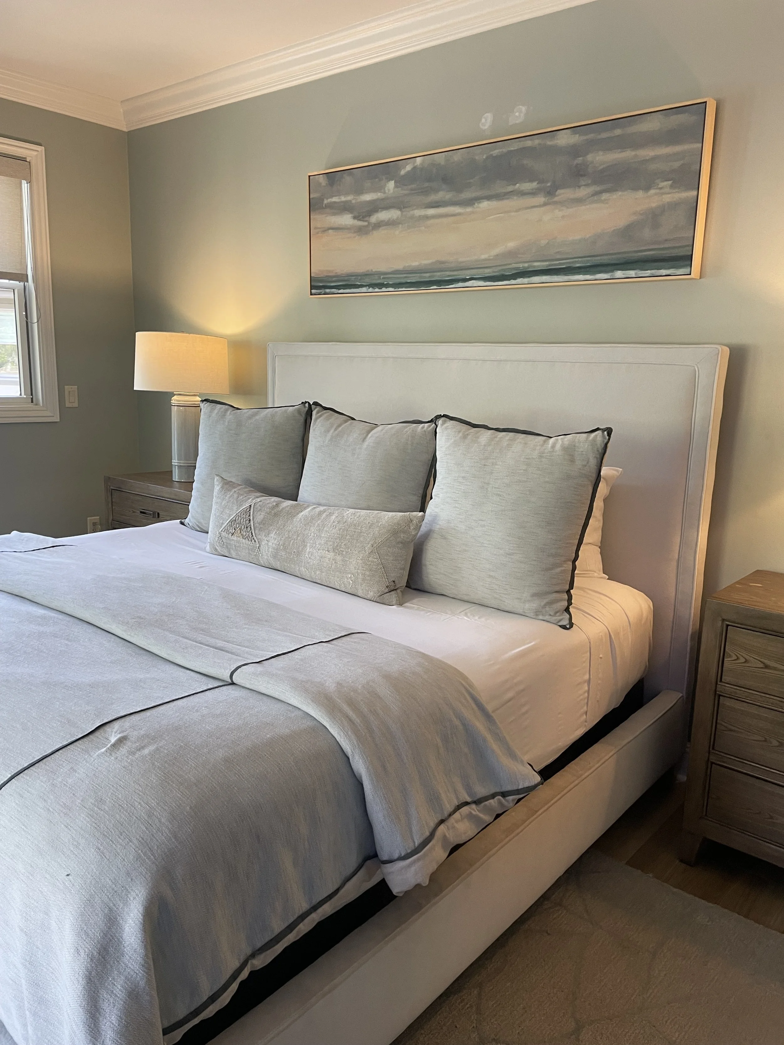 A neatly made bed with light-colored bedding and multiple pillows in a bedroom with mint green walls. There are two wooden nightstands with lamps on each side of the bed. A seascape painting hangs above the bed, and a window is visible on the left si