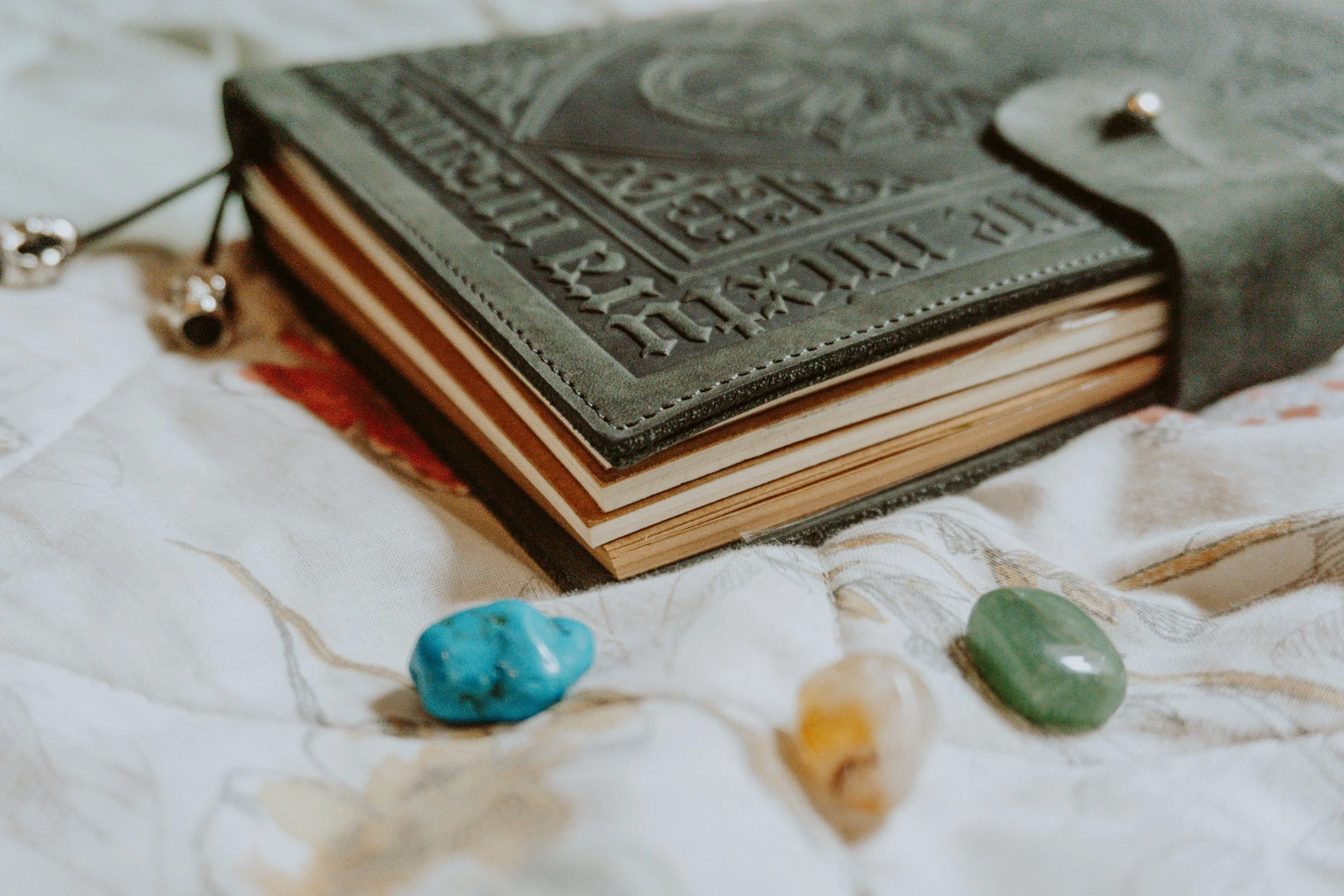 A black embossed leather-bound book or journal resting on a cloth surface with three small polished stones in front of it, one blue, one with yellow and white, and one green.