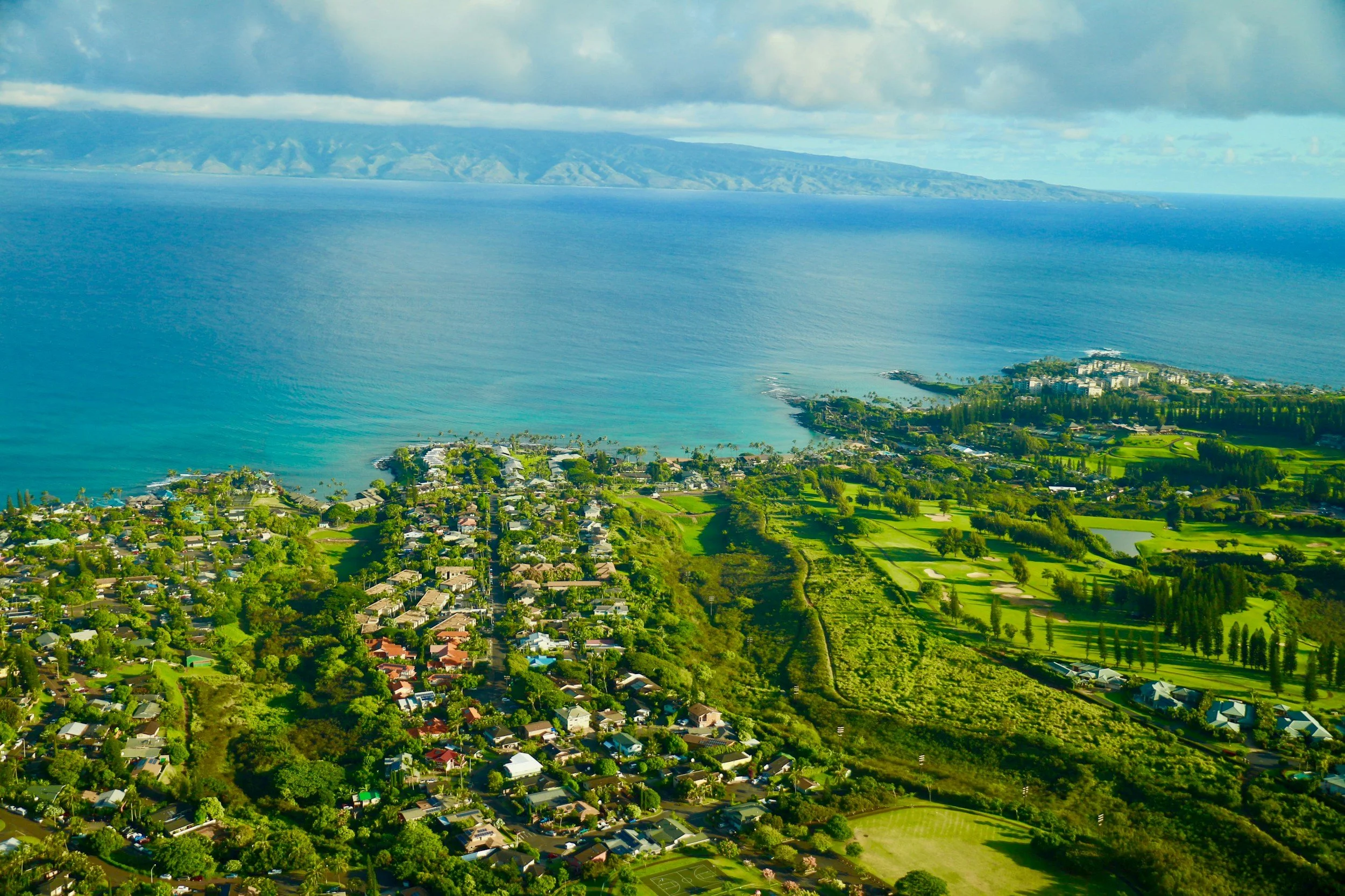 Aerial view of a coastal town with residential houses, green parks, and a golf course, along the shoreline with the ocean in the background, under a partly cloudy sky.
