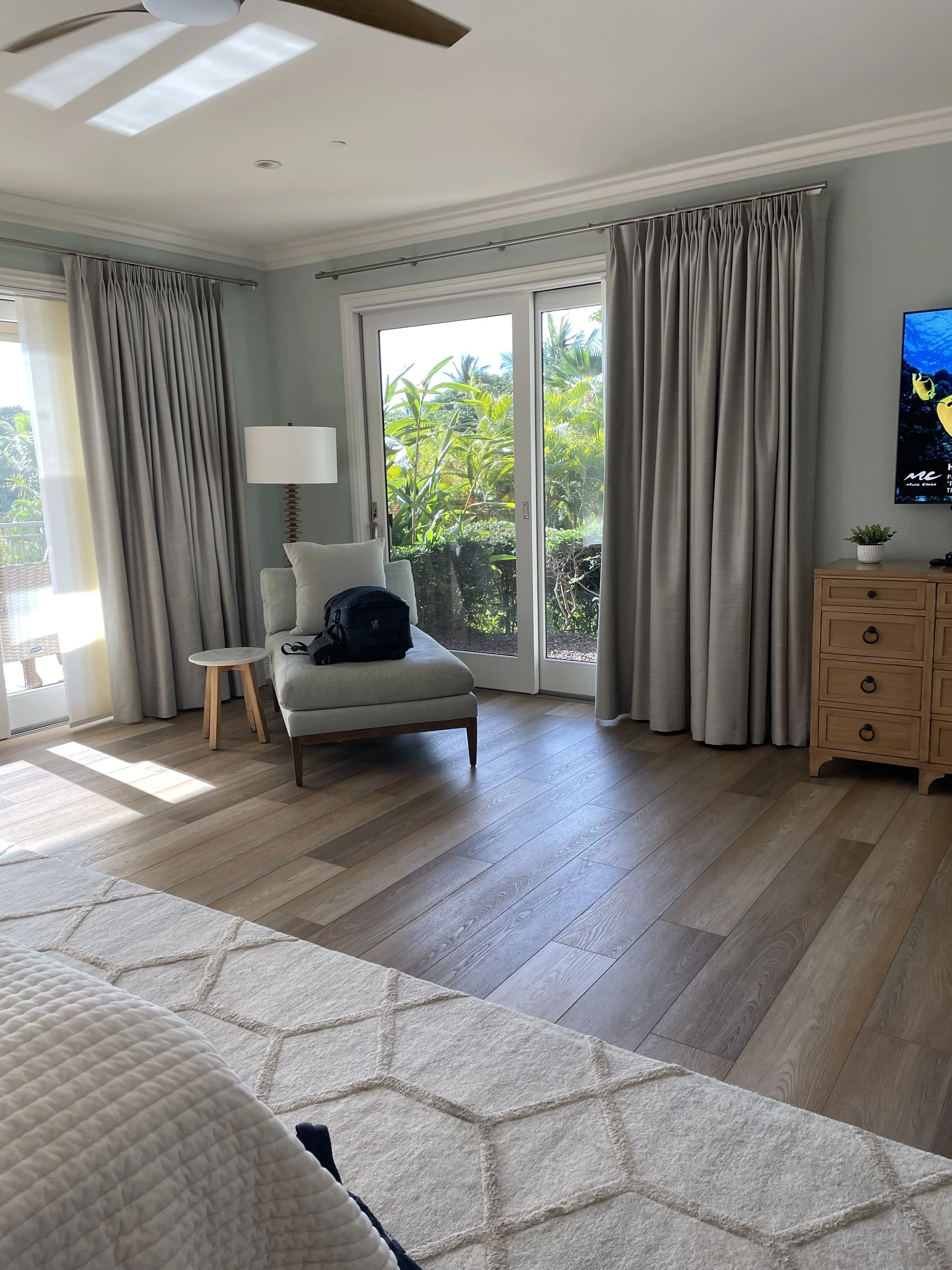 Bright living room with sliding glass door leading outside, beige curtains, a white armchair with a black bag on it, wooden side table, floor lamp, TV on wooden dresser, and green outdoor foliage visible through the door.