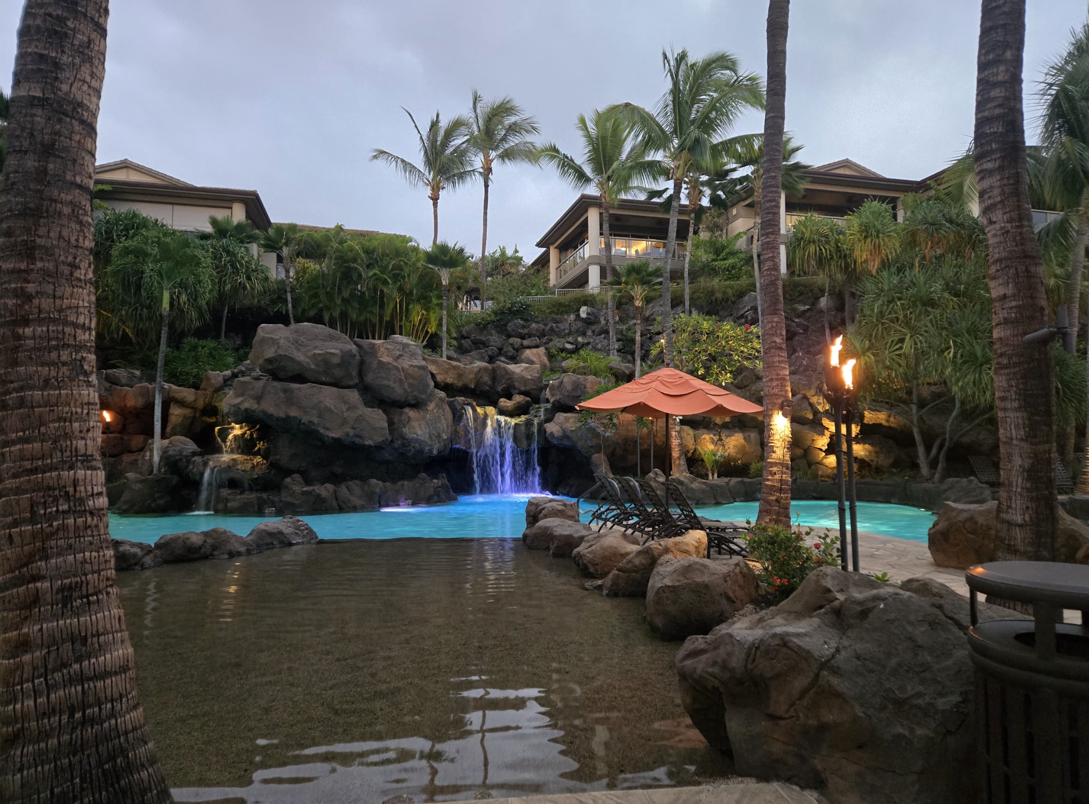 Resort pool area with a waterfall, palm trees, lounge chairs, an umbrella, and tiki torches at dusk.