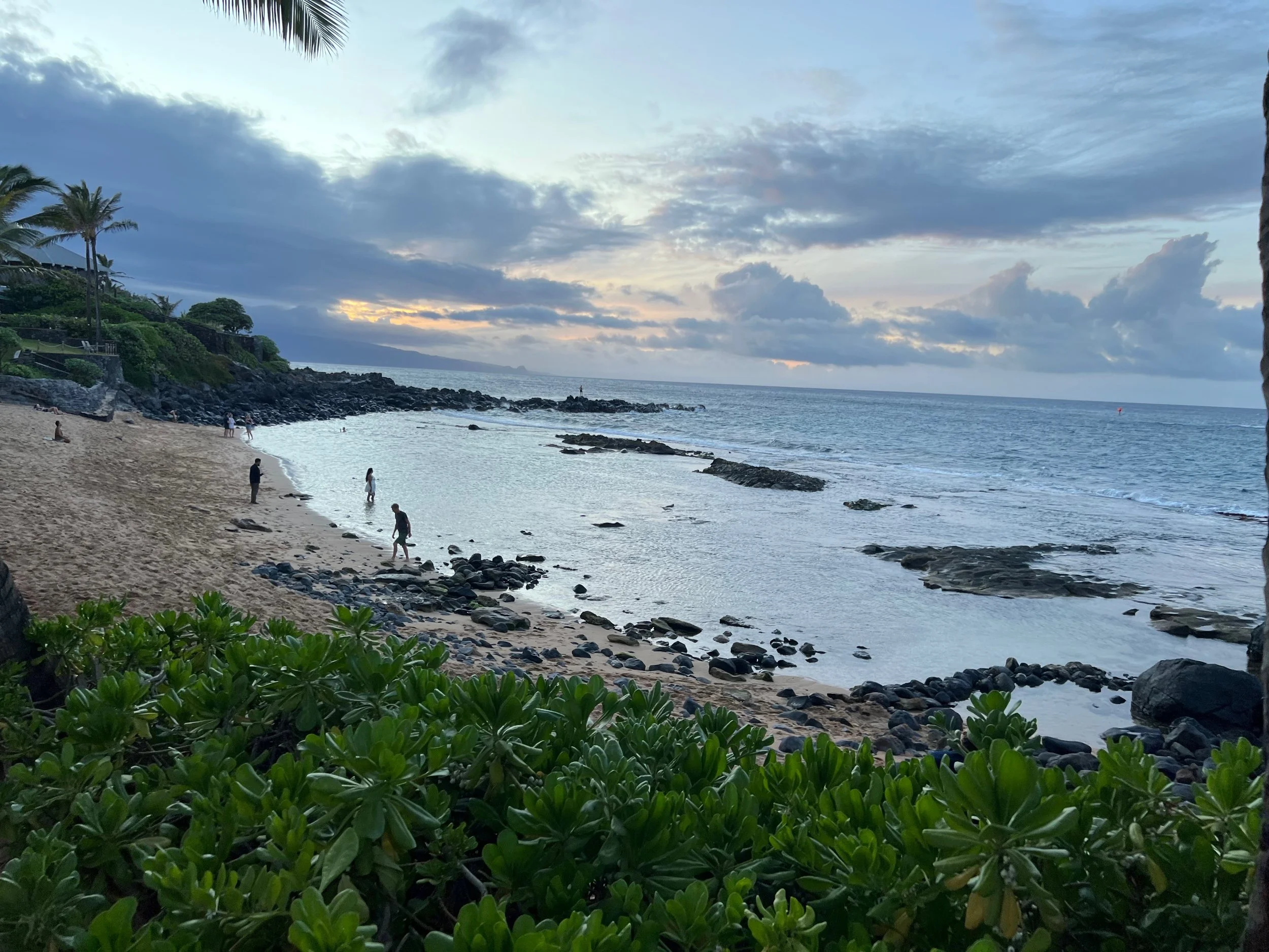 A tropical beach at sunset with a rocky shoreline, green foliage in the foreground, a few people walking on the sand, palm trees on a hill on the left, and a cloudy sky.