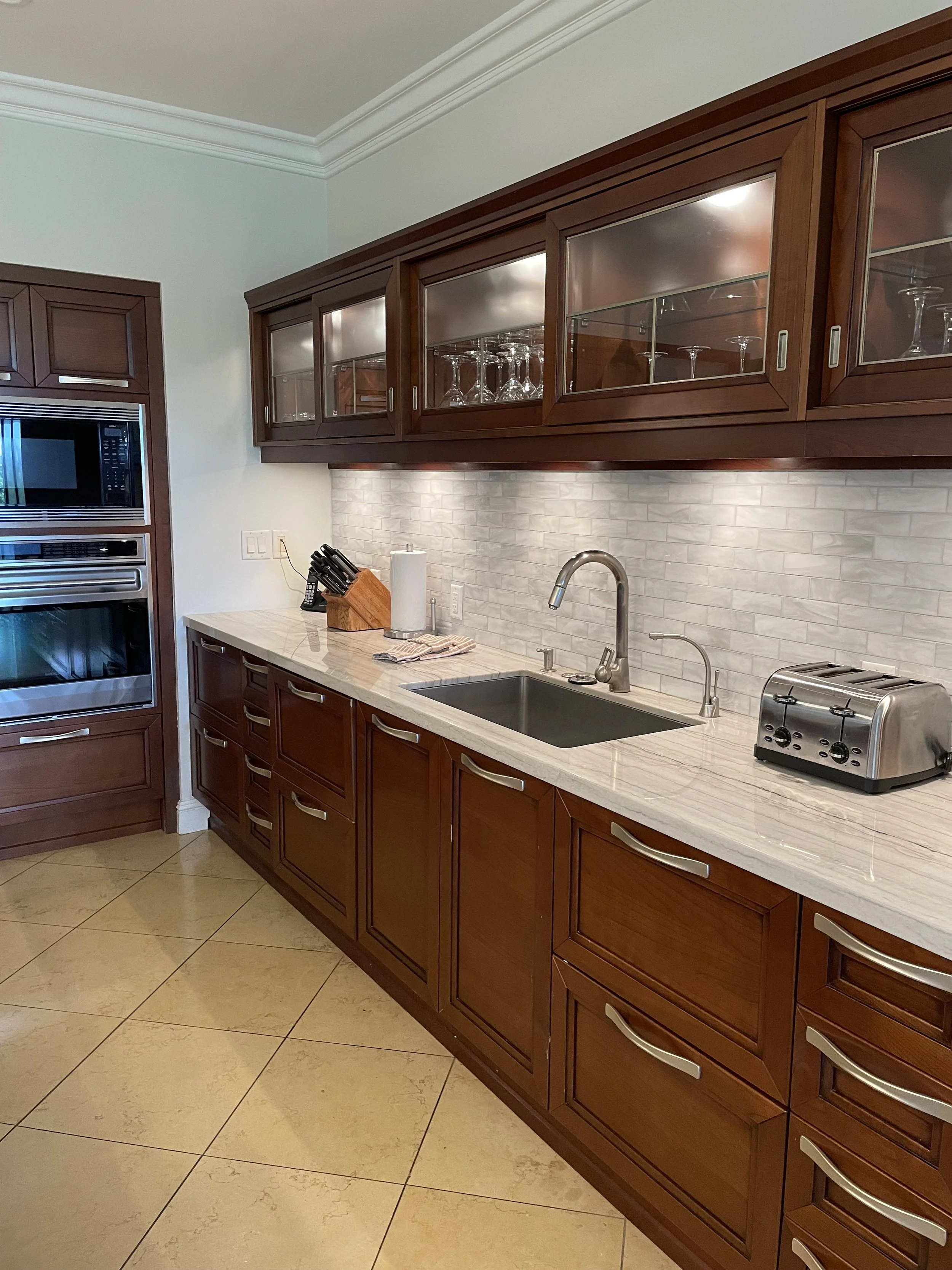 Kitchen with dark wooden cabinets, white tiled backsplash, and beige tile floor, featuring a sink, a toaster, and various utensils on the countertop.