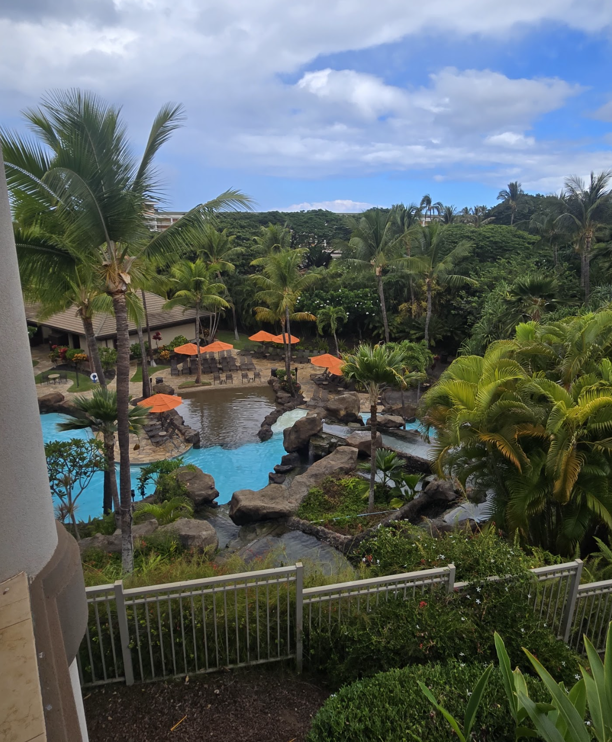 View of a tropical resort pool area surrounded by palm trees, with lounge chairs, orange umbrellas, and lush greenery under a partly cloudy sky.
