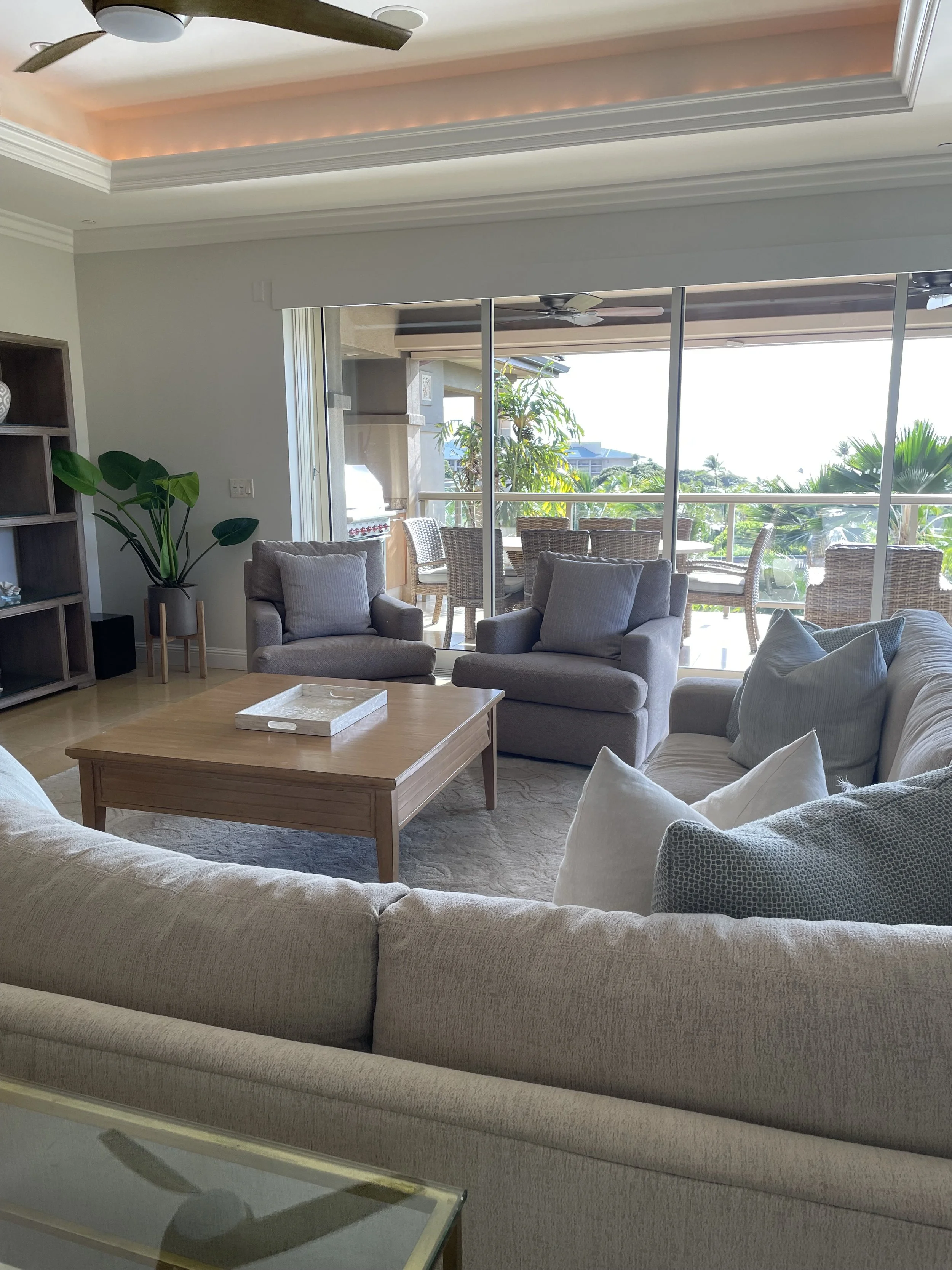 Living room with beige sofa, armchairs, wooden coffee table, large sliding glass doors leading to a balcony with outdoor dining table and plants, ceiling fan, shelves, and tropical view outside.