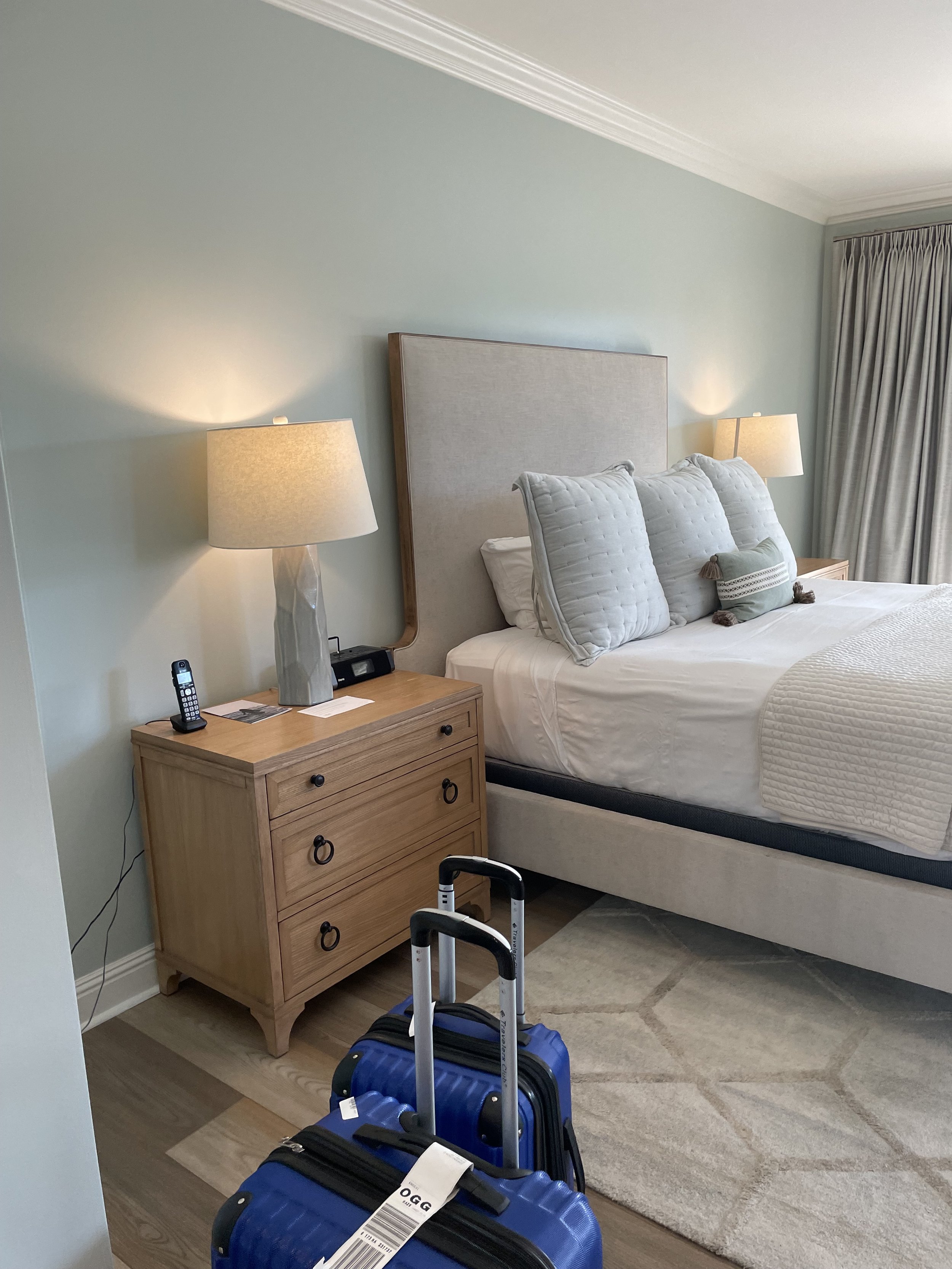 Hotel room with a large bed, nightstands with lamps, a lamp on the left nightstand, blue suitcases in the foreground, beige carpet, light green walls, and a window with beige curtains.