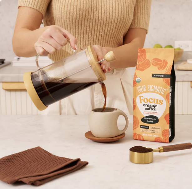 Person pouring coffee from a french press into a mug in a kitchen with a bag of Four Sigmatic Focus coffee and a wooden scoop on the counter.