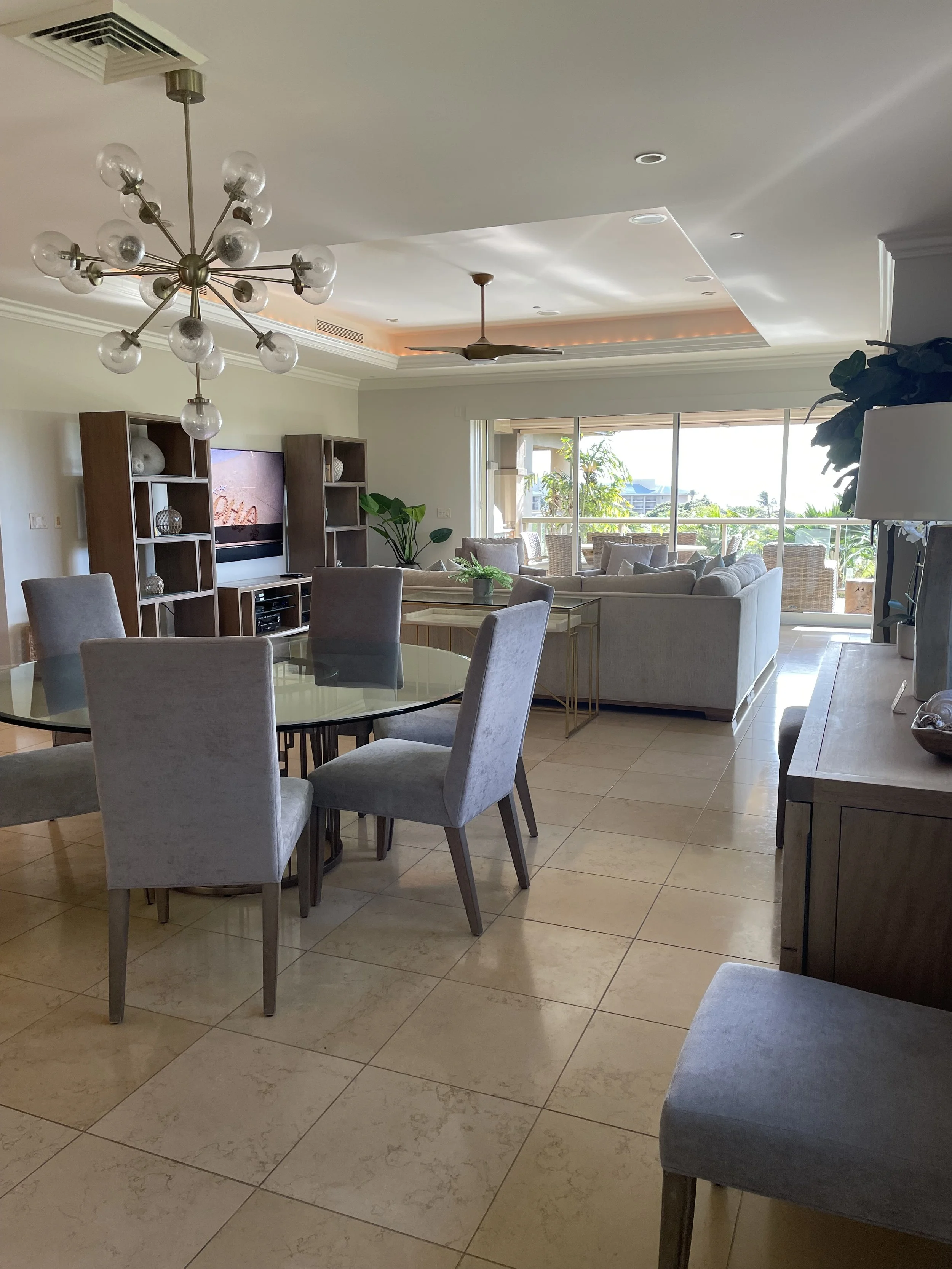 Living room with a dining table and six chairs, a sectional sofa, a TV with a console, large window with a balcony, plants, and ceiling lights.