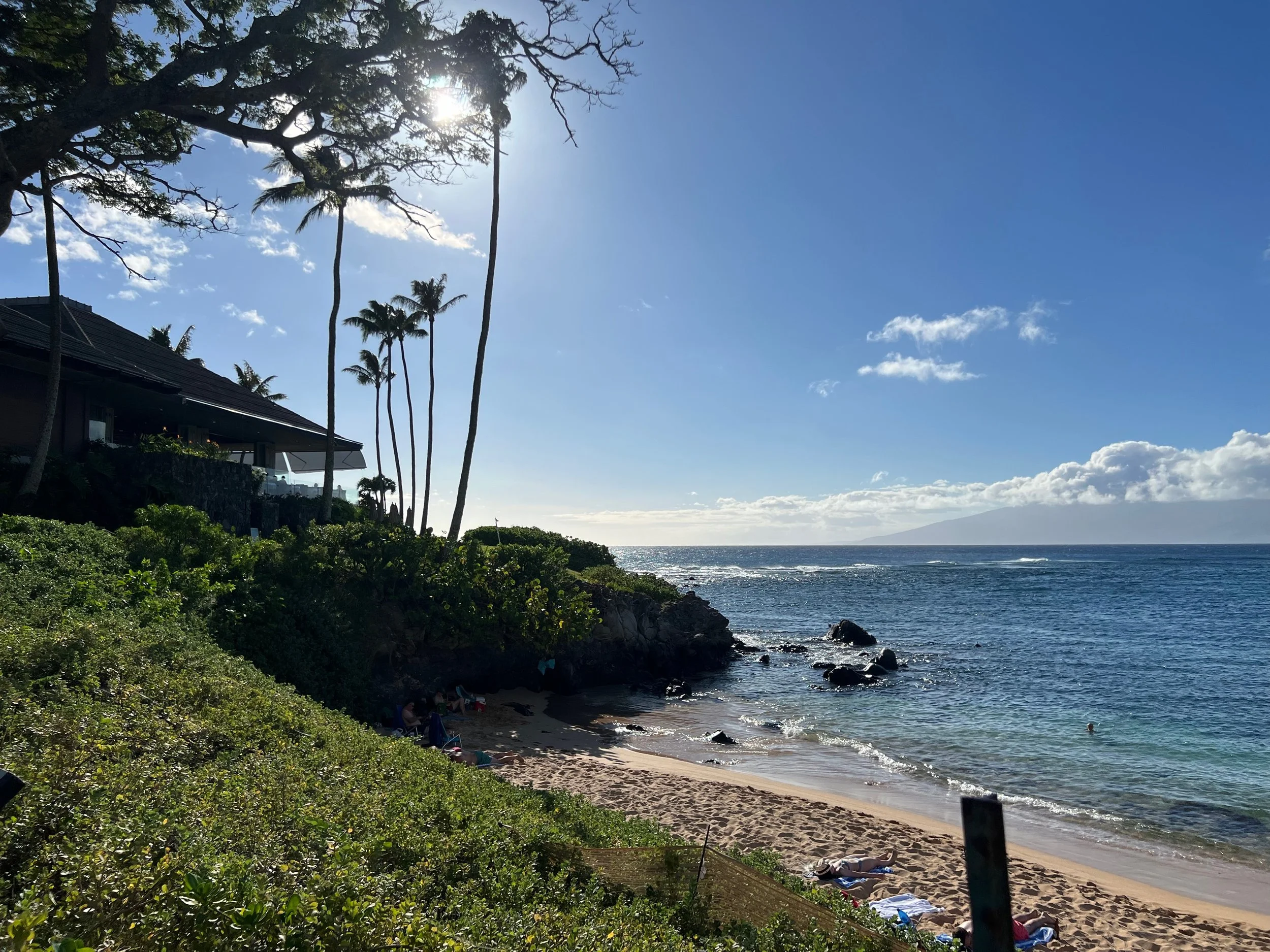Sun shining over a tropical beach with palm trees, a house on the left, and people relaxing on the sand, with the ocean and sky in the background.