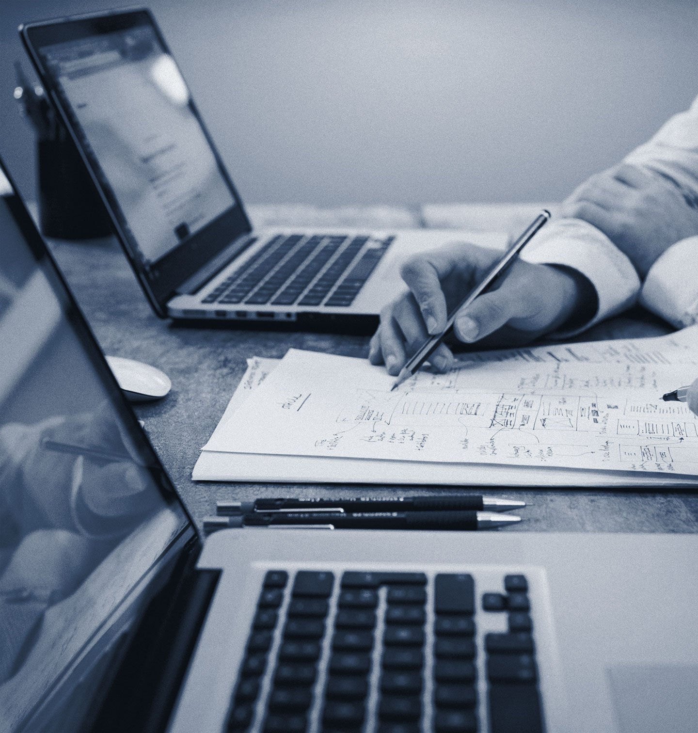 A person working at a desk with two laptops, writing on a sheet of paper with handwritten notes and diagrams, with pens nearby.