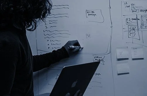 Person writing on a whiteboard with notes and diagrams, holding a marker in one hand and a laptop in the other.