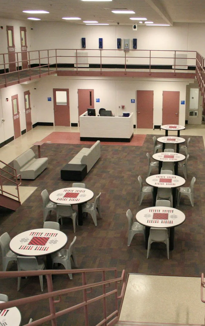 An empty indoor recreational or community center with round tables decorated with game-themed patterns, chairs, gray couches, and a reception desk. The space has a two-story layout with a balcony and a staircase visible in the foreground.