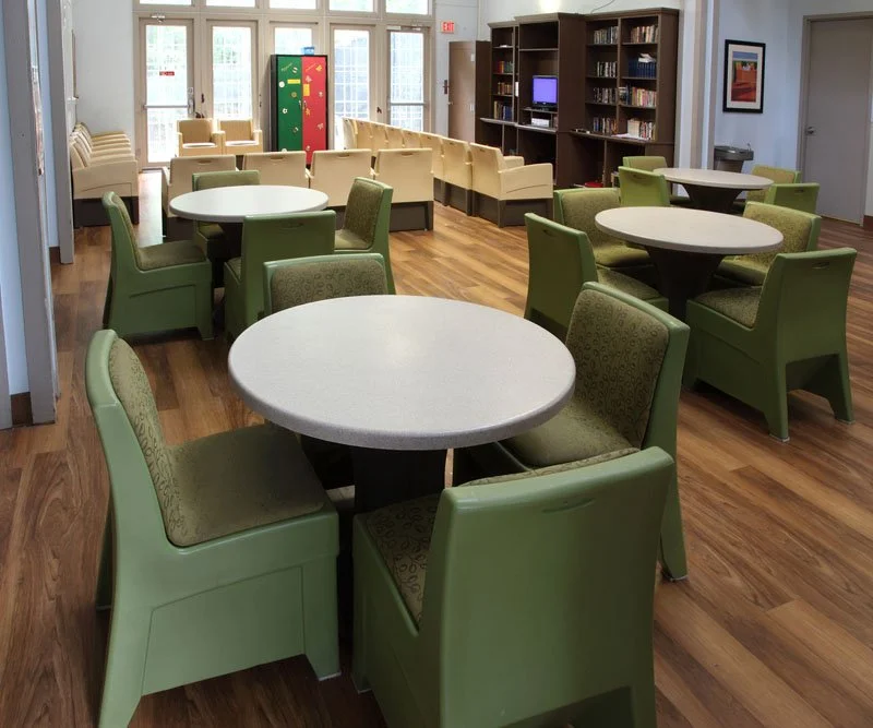 A spacious common room with green and beige chairs around white circular tables, a wooden floor, and a bookshelf along the wall. There is a windowed wall in the background, and a colorful locker or cabinet.