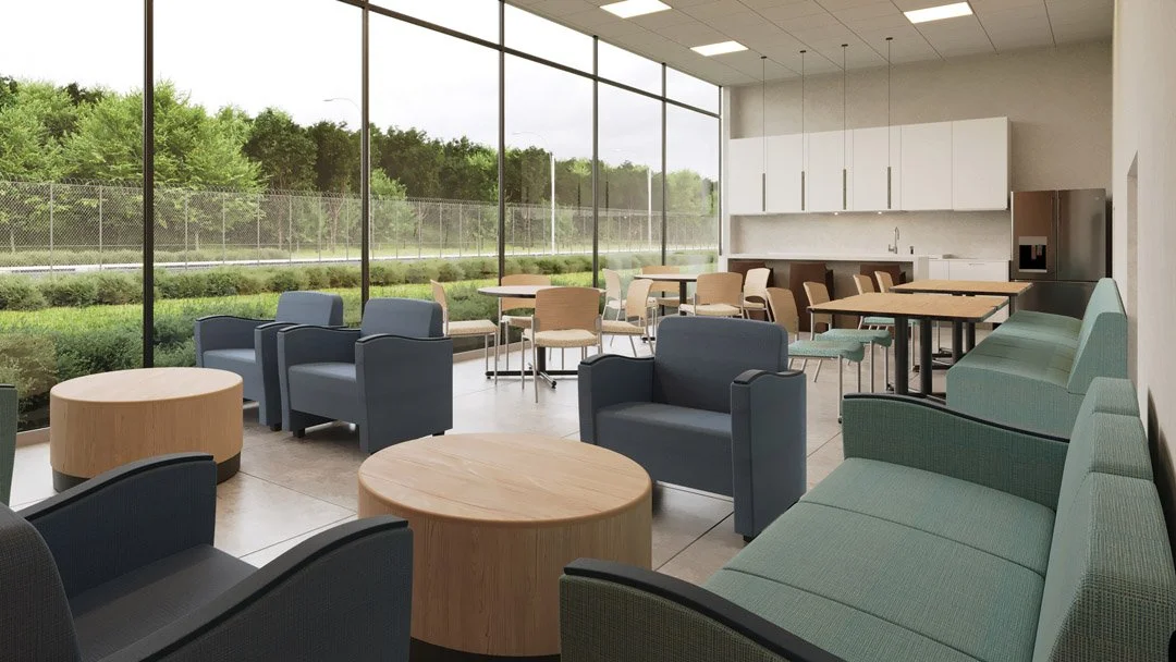 Indoor lounge area with blue and green armchairs, wooden coffee tables, and a green couch, featuring large floor-to-ceiling windows overlooking greenery and a fence outside.