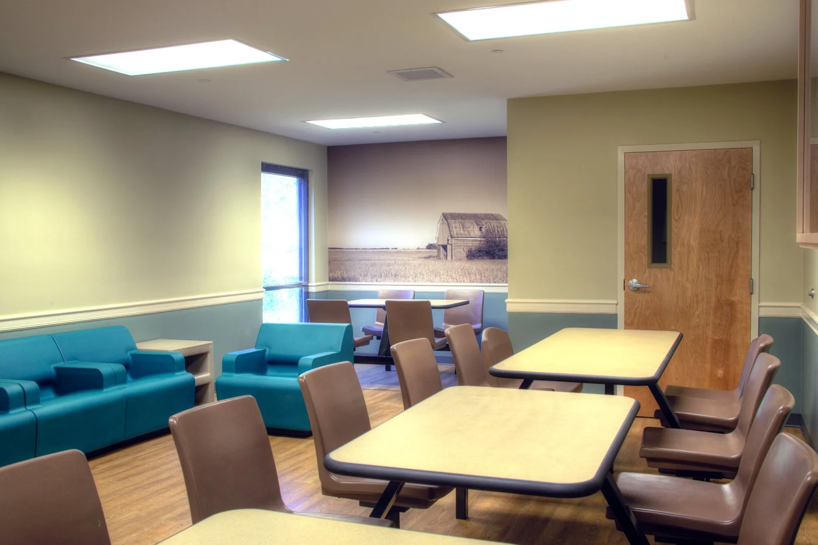 Indoor room with beige walls, teal sofa, brown chairs, wood flooring, and a large window with a farm barn mural.
