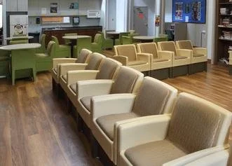 Empty waiting area with beige and green chairs arranged in rows in a room with wooden flooring.