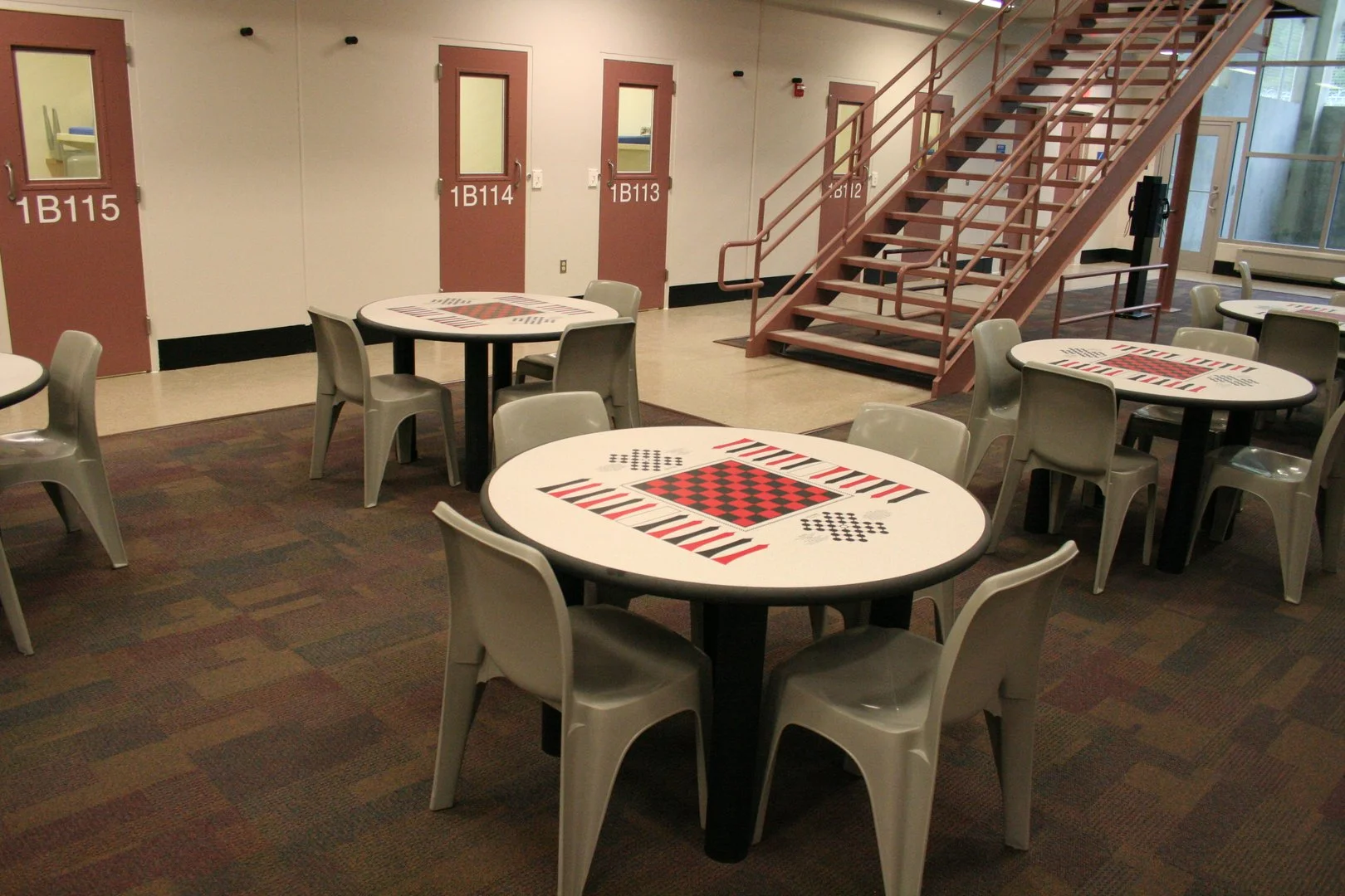 Indoor area with round tables featuring checkered patterns on the surface, gray chairs around the tables, beige walls with pink doors labeled 1B113, 1B114, 1B115, and a staircase with pink railing leading up to a second level, large windows to the right letting in natural light.