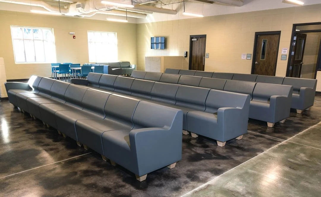 Empty seating area with rows of blue and gray chairs in a modern, well-lit room with beige walls, glossy dark floors, and large windows.