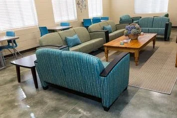 Waiting room with multiple sofas and chairs, a coffee table with a floral arrangement, and windows with blinds.