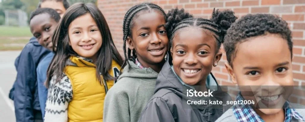 Five diverse children standing in line outdoors, smiling, with brick wall background.