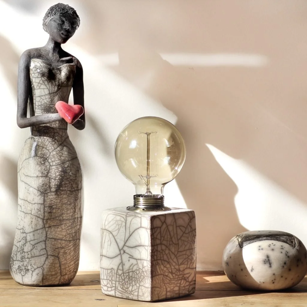 Statue de femme en céramique noire et blanche tenant un cœur rouge, lampe à incandescence vintage avec un globe en verre, et un récipient en céramique noir et blanc sur une surface en bois. Réalisé avec la technique "Raku"