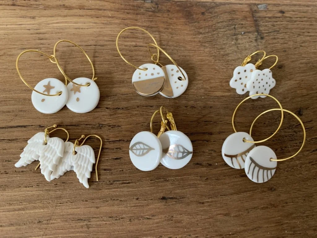 Ensemble de six paires de boucles d'oreilles en céramique de différentes formes avec motifs en or, posées sur un plan en bois.