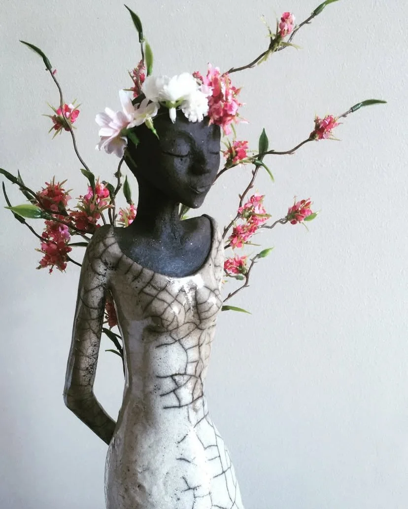 Statue de femme avec un visage serein, le corps en céramique craquelée, créer grâce à une technique de cuisson appeler raku, et une couronne de fleurs avec des branches fleuries
