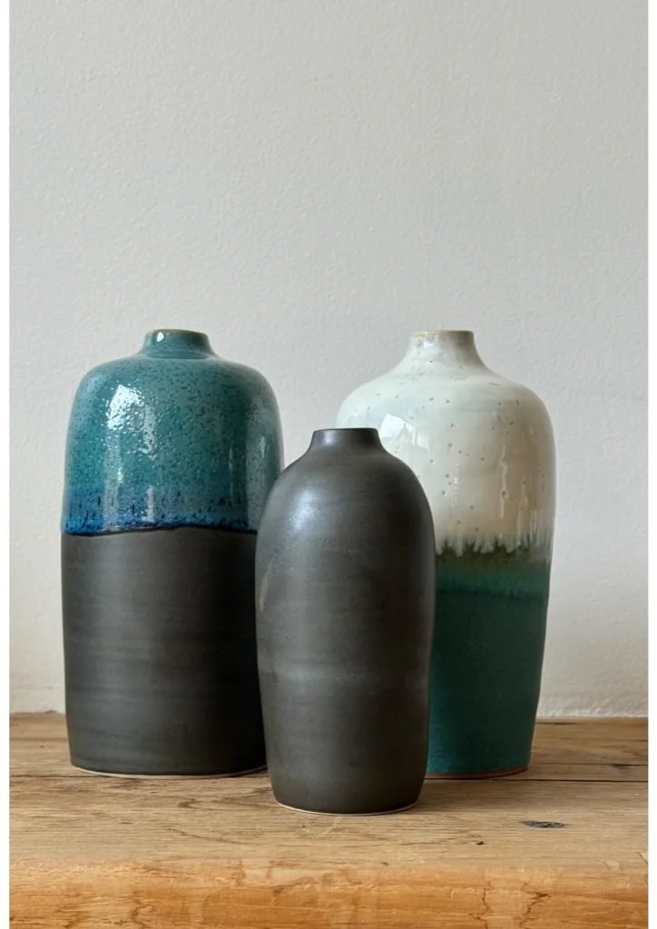 Trois vases en céramique de différentes tailles et couleurs, placés sur une table en bois, contre un mur blanc.