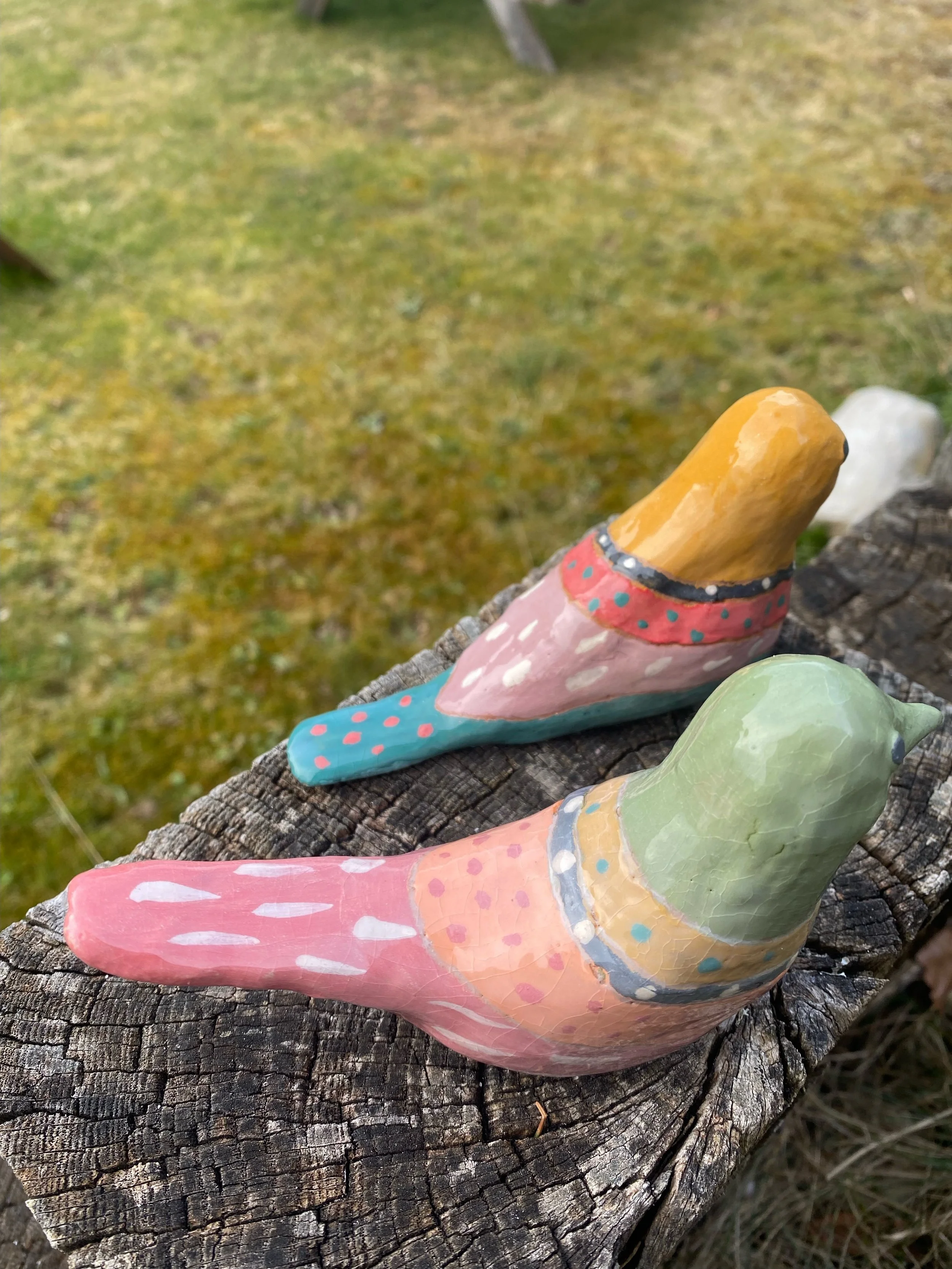 Deux oiseaux en céramique peint à la main, posés sur un vieux troncs d'arbre, avec une surface de sol recouverte de mousse et de feuilles en arrière-plan.