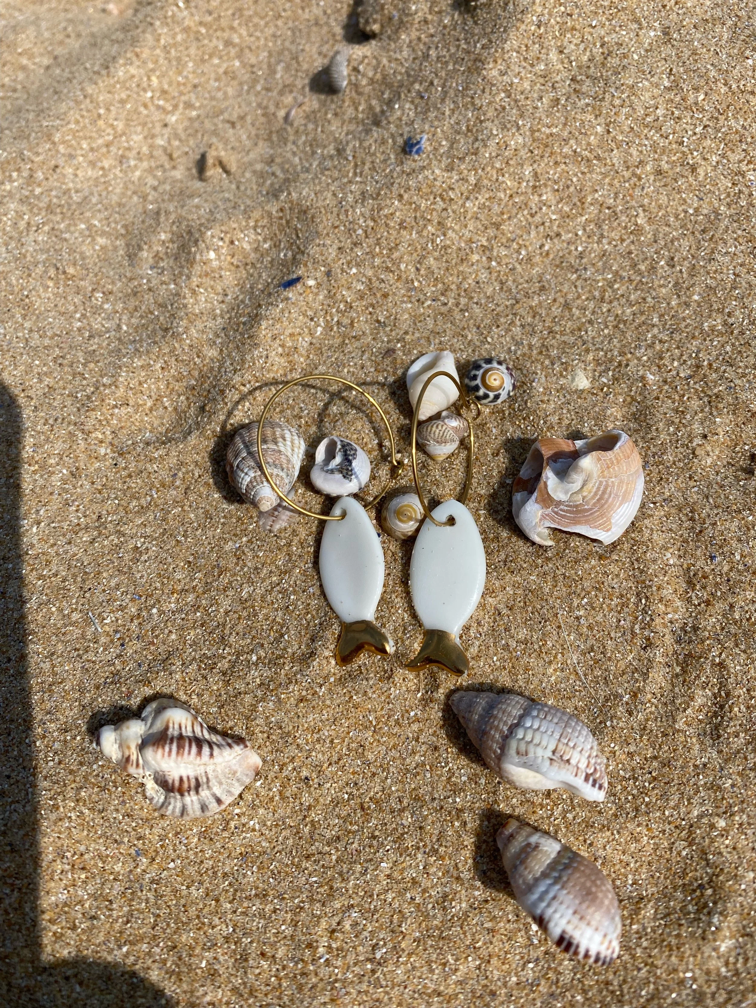 Une paire de boucles d'oreilles en forme de poisson posées sur du sable avec plusieurs coquillages autour.
