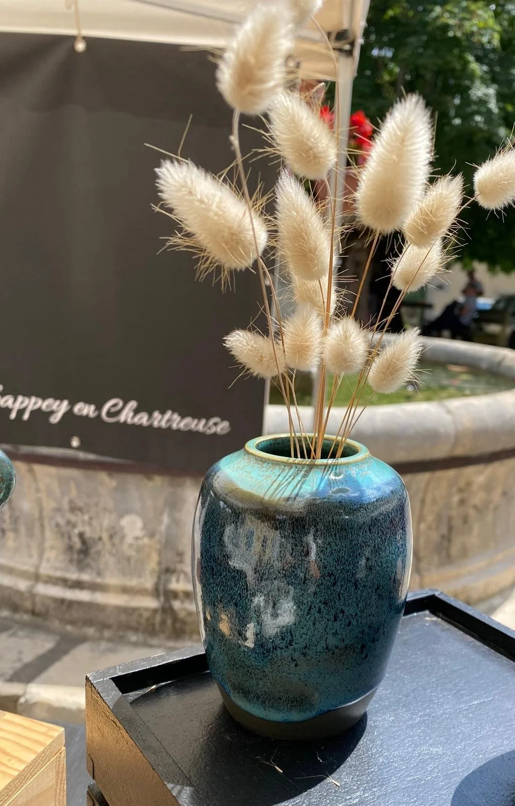 Un vase en céramique bleu et vert avec des tiges fleuries blanches, placé sur une surface noire à l'extérieur, avec un fond flou d'arbres et d'un banc.