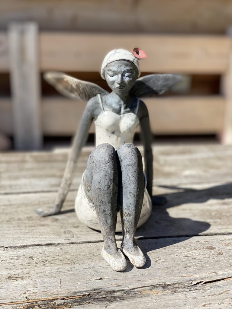Statue artisanale d'une fée assise avec un chapeau rose et des ailes en bois, placée sur une surface en bois extérieur.