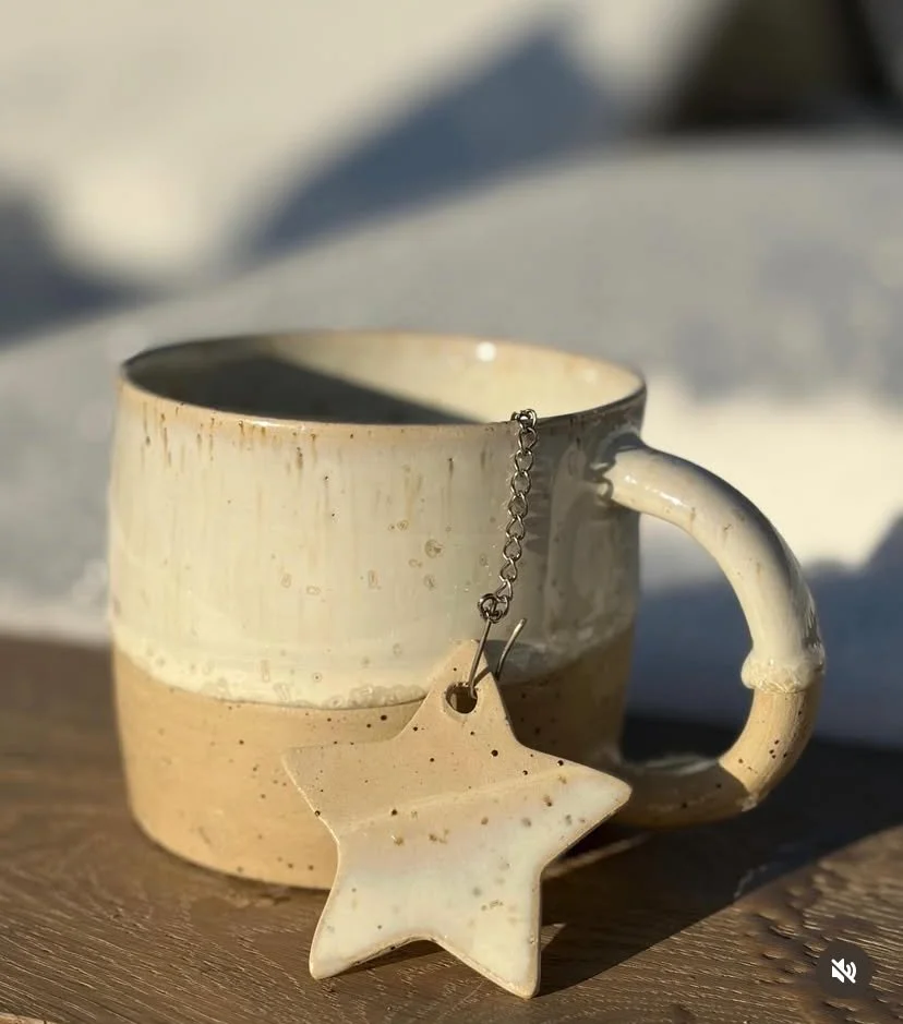 Tasse en céramique beige avec une étoile en porcelaine suspendue par une chaîne, posée sur une surface en bois. Fond flou avec lumière naturelle